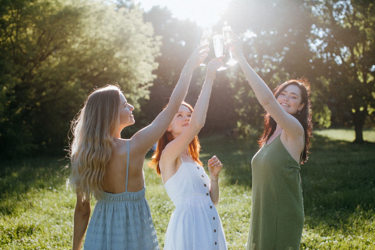 Three women toasting with drinks in a scenic summer field.