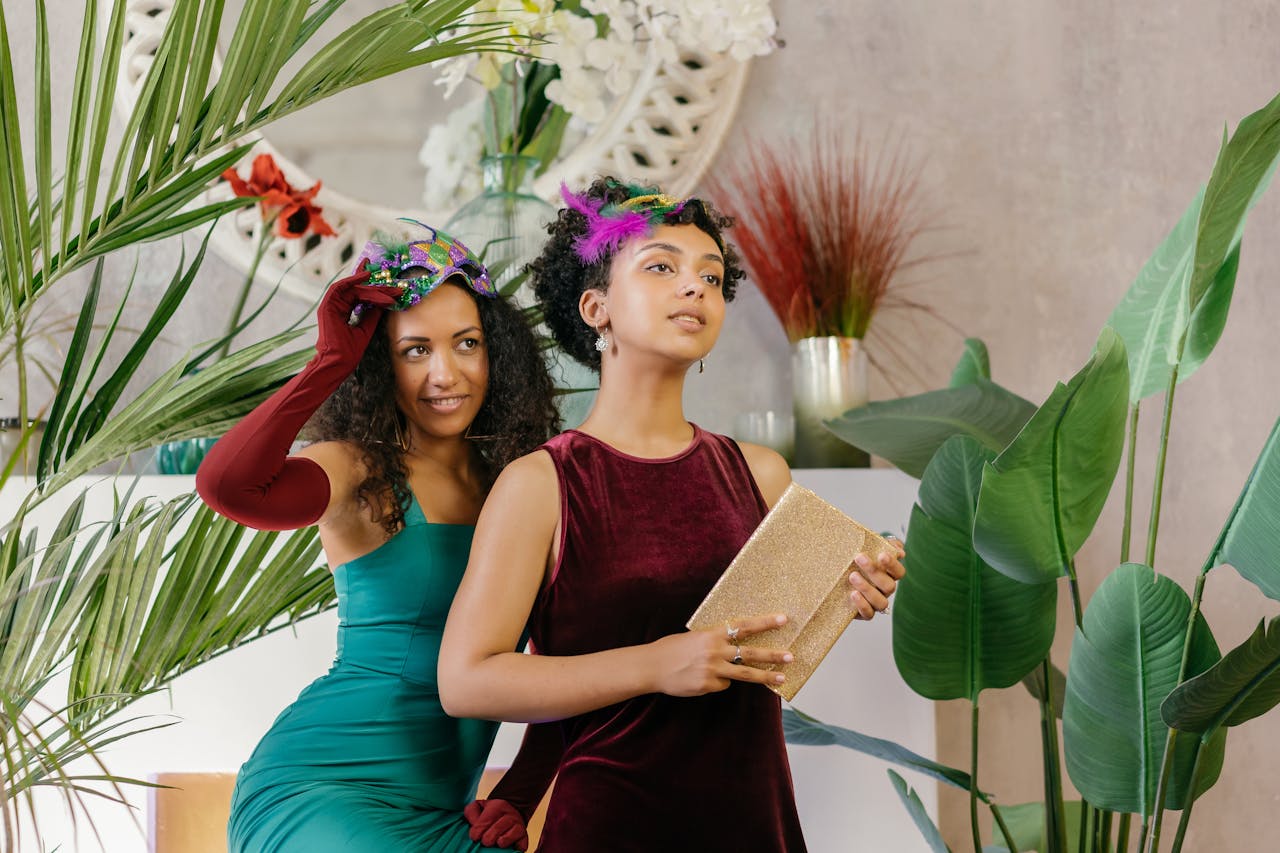 Two women in elegant dresses and masks pose stylishly at an indoor masquerade party setting.